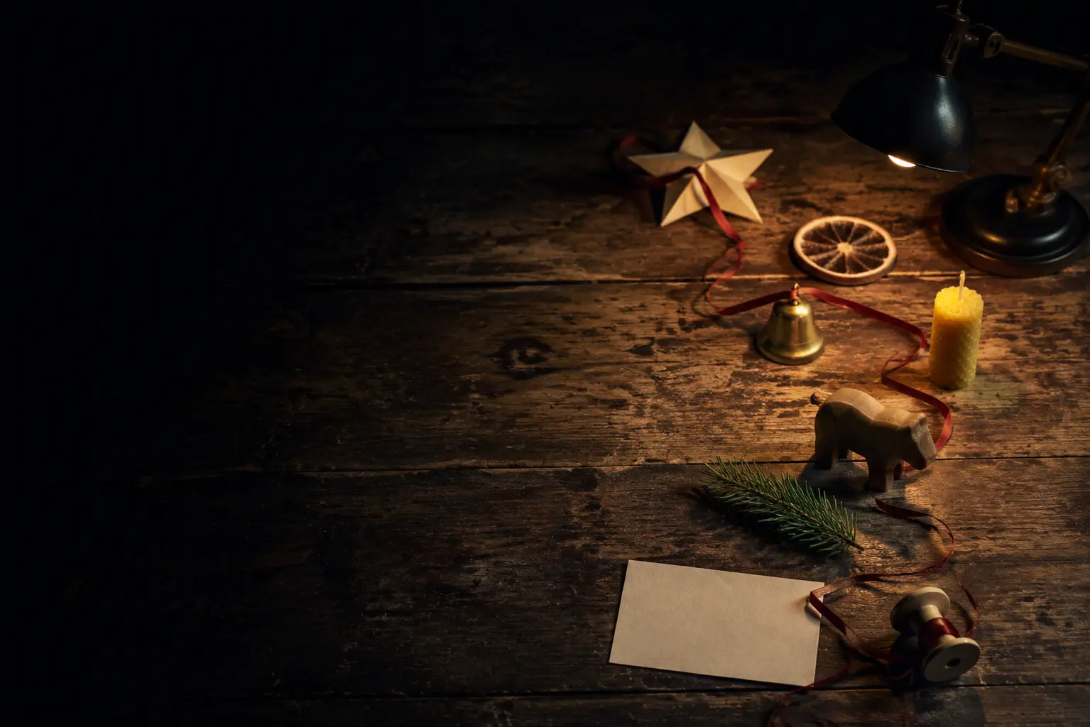 Small real handmade holiday objects connected by one narrow red ribbon on a worn wooden worktable before dawn
